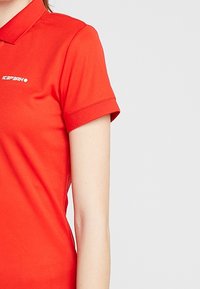Lyhythempparinen punainen polo paita, joka on valmistettu sileästä materiaalista. Paidassa on nappikaulus ja valkoinen logo vasemmassa rinnassa.