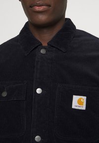 Giacca di velluto a coste blu navy con colletto, bottoni a pressione, due tasche frontali e una toppa rettangolare con il logo Carhartt. Dettagli del tessuto testurizzato visibili.