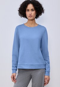 Femme aux cheveux bouclés portant un sweat-shirt bleu clair à manches longues et un pantalon gris à fines rayures, debout devant un fond uni.
