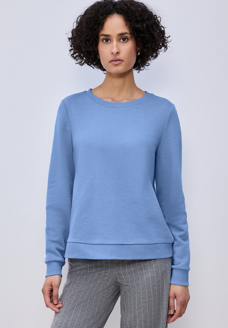Femme aux cheveux bouclés portant un sweat-shirt bleu clair à manches longues et un pantalon gris à fines rayures, debout devant un fond uni.