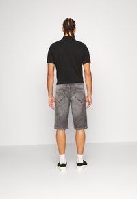 Mies joka on punotulla hiuksella, päällä musta polo-paita, harmaat polvimittaiset farkkushortsit, valkoiset sukat ja mustat lenkkarit, seisoo selin.