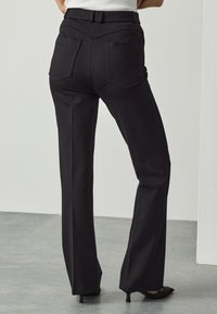 Black trousers with a tailored fit, featuring a high waist, two back pockets, and wide legs. Made from a textured fabric with minimal detailing.