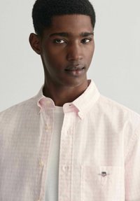 Camicia button-up a quadri rosa chiaro realizzata in cotone, con taschino frontale con dettaglio del logo e colletto classico, indossata sopra una maglietta bianca.