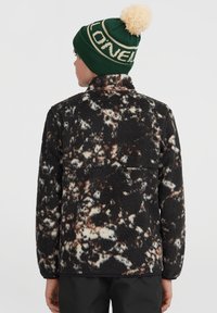Giacca in pile a motivi neri e beige con colletto alto. Un berretto lavorato a maglia verde con pompon beige completa l'outfit.