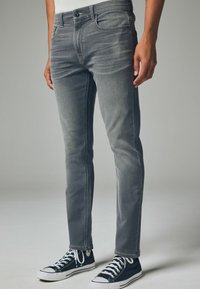 Blugi slim-fit gri cu accente ușor distresate, buzunare frontale și închidere cu nasture metalic, asortați cu adidași negri și albi.