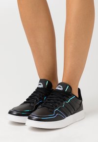 Svart sneaker med glänsande detaljer, vit sula, texturerad ovandel och flerfärgad kant. Har svarta snörningar och logotyp på tungan.