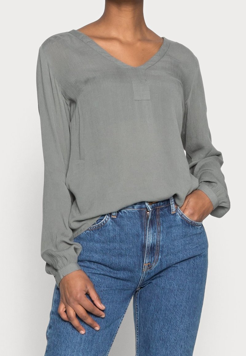 Femme portant un chemisier gris à manches longues, ample et col en V, rentré dans un jean bleu taille haute, une main dans la poche.