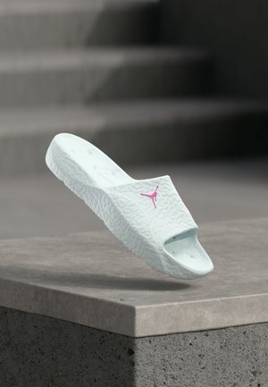 Λευκό σαγιονάρι με υφή, με ροζ λογότυπο Jumpman να αιωρείται πάνω από μια τσιμεντένια άκρη με θολές σκάλες στο φόντο.