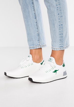 Persona che indossa scarpe da ginnastica bianche con lacci con logo di canguro verde e jeans azzurri, in piedi su un pavimento bianco.