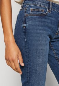 Gros plan d'une main d'une personne reposant à côté d'un jean en denim bleu avec des coutures orange et une petite poche avant.