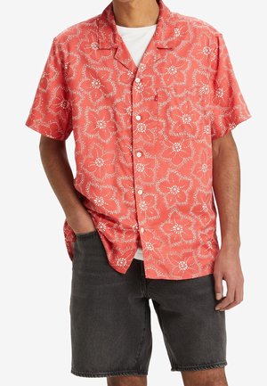 Mann trägt ein korallenfarbenes kurzärmliges Hemd mit weißen Blumenmustern über einem weißen T-Shirt, kombiniert mit schwarzen Jeansshorts, eine Hand in der Tasche.