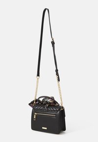Sac en cuir noir à bandoulière avec texture matelassée, accents en chaîne dorée, et un foulard à motifs colorés noué sur le dessus. Poche zippée à l'avant.
