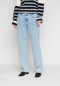 Vaqueros de mezclilla azul claro con un corte recto. Presentan cinco bolsillos, pases para cinturón y un ligero desgaste. Combinados con un suéter a rayas en blanco y negro.