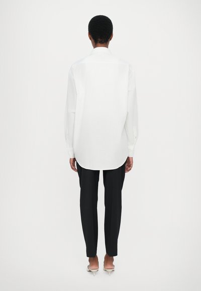 Long-sleeve white blouse with a curved hem, viewed from the back, paired with black fitted trousers and pointed toe heels on a neutral background.