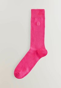 Svetlo roza bombažna nogavica z rebrastim robom, gladko teksturo in prilegajočo obliko. Ima majhen logo blizu roba.