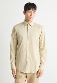 Cămașă bej deschis cu mânecă lungă, cu guler tip button-down, textură netedă și o ușoară strălucire, având un tiv curbat și nasturi albi.