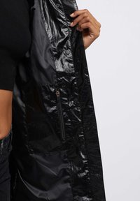 Cappotto impermeabile nero con una finitura lucida, dotato di tasca con zip, chiusure a pressione e una vestibilità comoda. Materiale testurizzato visibile.