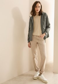 Giacca grigia chiara con cappuccio sopra un maglione beige, abbinata a pantaloni beige e sneakers bianche. Texture morbide e palette di colori neutri.