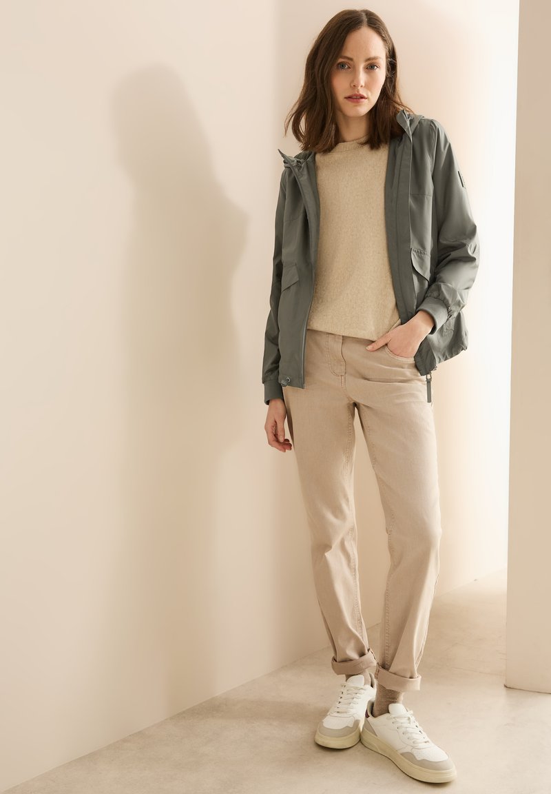 Giacca grigia chiara con cappuccio sopra un maglione beige, abbinata a pantaloni beige e sneakers bianche. Texture morbide e palette di colori neutri.