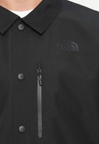 Pogled od blizu na črno jakno North Face, ki prikazuje ovratnik, blagovne znamke gumbke na zapenjanju, žep na zadrgo na prsih in izvezeno logo.