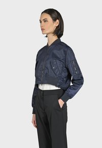 Marinblå kort bomberjacka med dragkedja fram, ribbade ärmslut och fickor; slät, glänsande tyg med detaljer av sidofickor med dragkedja.