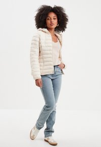 Giubbotto imbottito avorio con cappuccio, chiusura con zip e texture trapuntata. Abbinato a jeans azzurri e scarpe da ginnastica bianche.