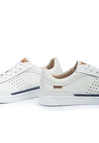 Zapatillas de piel blanca con diseño perforado, acentos en color beige y cordones blancos. Presentan una franja azul oscuro en contraste y una etiqueta con el logo.