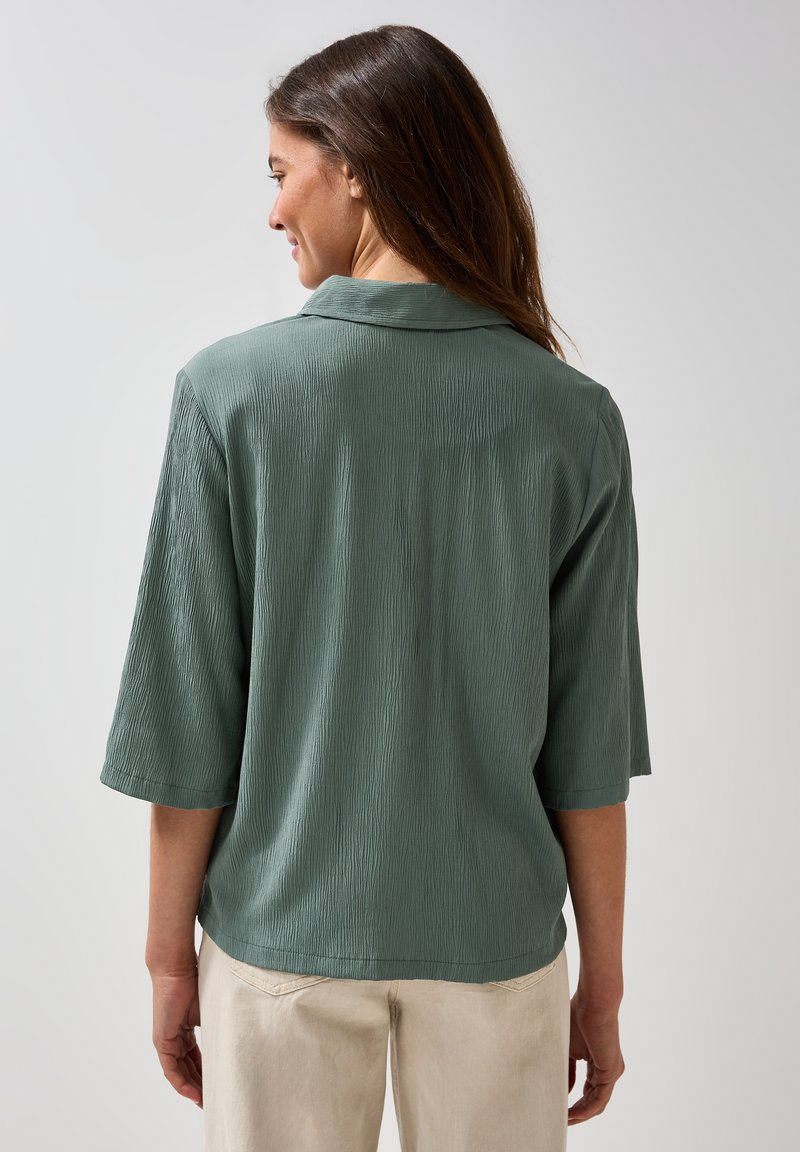 Blusa verde, con texture, a maniche corte e colletto. Vista da dietro, abbinata a pantaloni di colore chiaro.