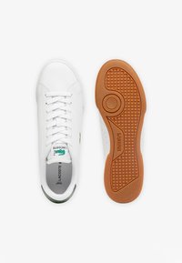 Witte leren sneakers met groene accenten, ronde neus, geperforeerde zijkanten, platte witte veters en een textuur rubberen zool. Lacoste-logo zichtbaar.