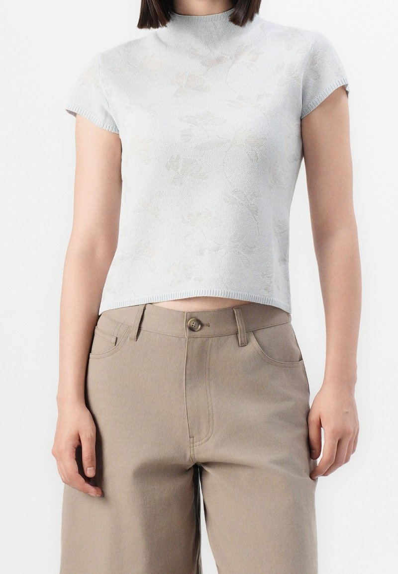 Femme portant un haut en maille à manches courtes à fleurs gris clair, légèrement rentré sur un pantalon beige taille haute avec boutons et poches.