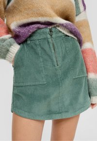 Falda de pana verde con una cinturilla elástica, bolsillo trasero con cremallera y dos bolsillos frontales. Presenta un diseño de textura acanalada.
