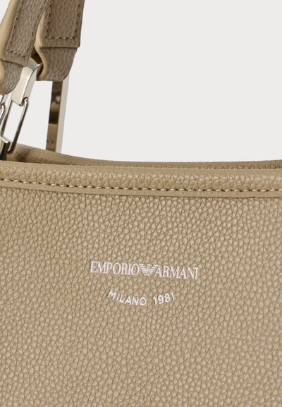 Τσάντα χειρός από μπεζ δερματίνη με υφή και καμπυλωτό σχέδιο, με ασημένιο λογότυπο και ανάγλυφο το όνομα της μάρκας. Ο ιμάντας είναι ρυθμιζόμενος και διακοσμημένος με λεπτομέρειες.