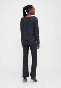 Svart perlebrodert langermet topp med en teksturert blomsterdesign, kombinert med svarte slengbukser og blanke svarte hæler.