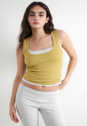 Jonge vrouw met lang donker haar die een gelaagde gele tanktop en grijze trainingsbroek draagt, staat voor een effen witte achtergrond.