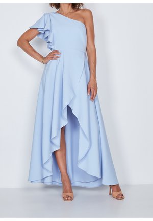 Kobieta w jasnoniebieskiej sukience na jedno ramię z falbaną na rękawie i asymetrycznym dołem, stojąca z jedną ręką na biodrze i ubrana w beżowe buty na obcasie z odkrytymi palcami.