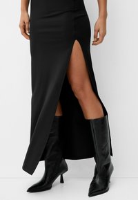 Svart satinmaxiklänning med hög slits, ihopparad med knähöga svarta stövlar med spetsig tå i texturerat läder.