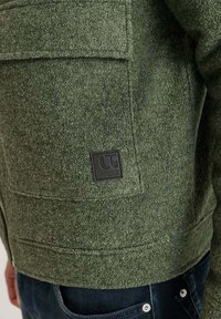 Chaqueta verde de fieltro con un bolsillo de parche y una etiqueta de logo negra. La tela tiene un acabado texturizado y cuenta con un cierre de cremallera. Jeans azul oscuro visibles.