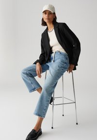 Zwarte blazer, crèmekleurige top met een zwarte rand, lichtblauwe wijde jeans en zwarte loafers, aangevuld met een lichtgrijze pet.