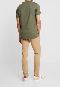 Uomo in piedi di spalle, che indossa una camicia a maniche corte verde oliva, pantaloni slim-fit beige e sneaker beige e bianche su uno sfondo neutro.