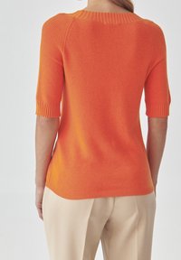 Donna che indossa un top aderente arancione a maniche corte in maglia e pantaloni beige, mostrata di spalle su uno sfondo semplice.