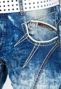 Blauwe denim jeans met lichtere verweerde delen, benadrukt door contrasterende bruine stiksels en een rood logo-label nabij de zak. Witte geperforeerde riem.