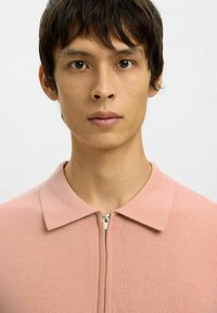 Jeune personne aux cheveux courts et foncés, portant un haut zippé rose pâle avec un col, faisant face vers l'avant sur un fond clair uni.