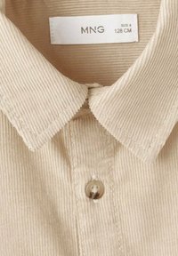 Beige Cordhemd mit spitzem Kragen, einem einzelnen Knopf am Hals und einer ausgeprägten vertikalen Rippstruktur. Markenlabel sichtbar.