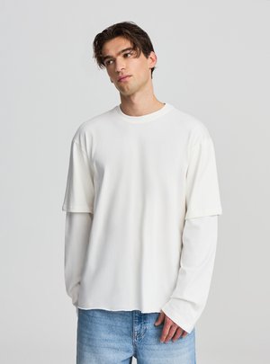 Maglietta bianca a maniche lunghe con strati, collo tondo e vestibilità rilassata, realizzata in tessuto liscio. Abbinata a jeans di denim azzurro chiaro.