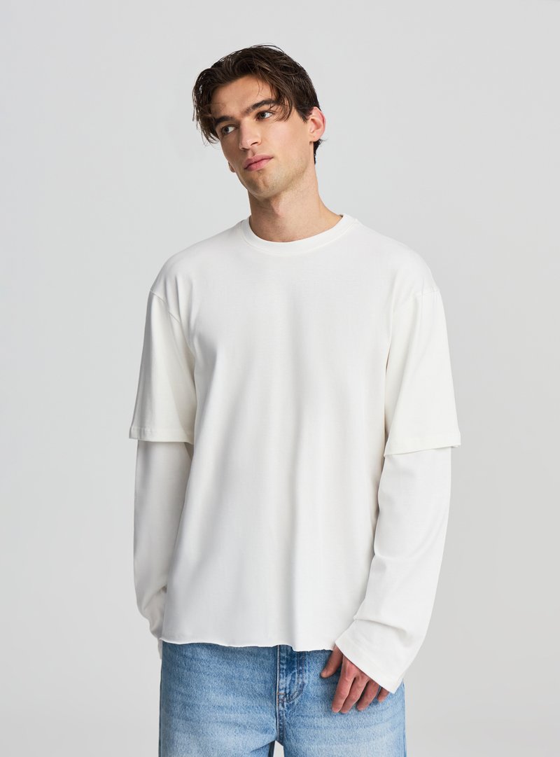 Maglietta bianca a maniche lunghe con strati, collo tondo e vestibilità rilassata, realizzata in tessuto liscio. Abbinata a jeans di denim azzurro chiaro.