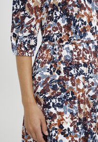 Blumendruckkleid mit Dreiviertelarm, das eine Krawatten-tailie hat. Das Material hat eine gekräuselte Textur in Marineblau, Braun und Hellblau auf einem weißen Hintergrund.