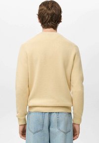 Żółty dziany sweter z prążkowanymi mankietami i ściągaczem u dołu, z klasycznym okrągłym dekoltem. Nosi się go na jasnoniebieskie szorty jeansowe. Widok z tyłu.
