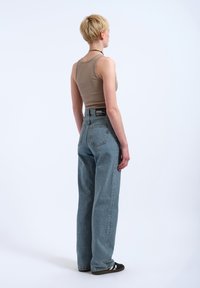 Frau mit kurzen blonden Haaren, die ein beiges Tanktop, lockere blaue Jeans mit hohem Bund und schwarze Turnschuhe trägt, steht mit dem Rücken zur Kamera.