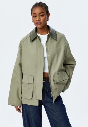 Green canvas jacket with a black leather collar, front pockets, and a zipped closure, paired with a white top and dark blue jeans.