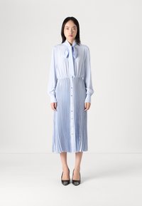 Vestido plissado azul claro com botões, mangas longas e detalhe de laço no pescoço. Combinado com sapatilhas pretas, com uma textura suave e um design fluido.